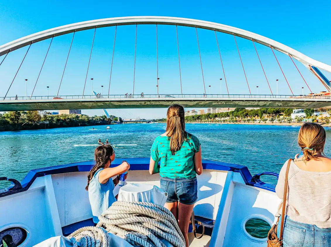 Passengers on the bow of a Seville boat tour approaching the white arch of the Barqueta Bridge